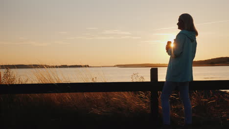 lonely woman drinks beer from a can on the lake standing alone by the fence looking at the sunset