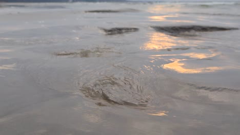 close up low angle footage of water flowing and swirling on sandy beach surface on a shoreline with sun glaring and reflecting on the wet sand in slow motion. ocean sea water waves visibe at back.
