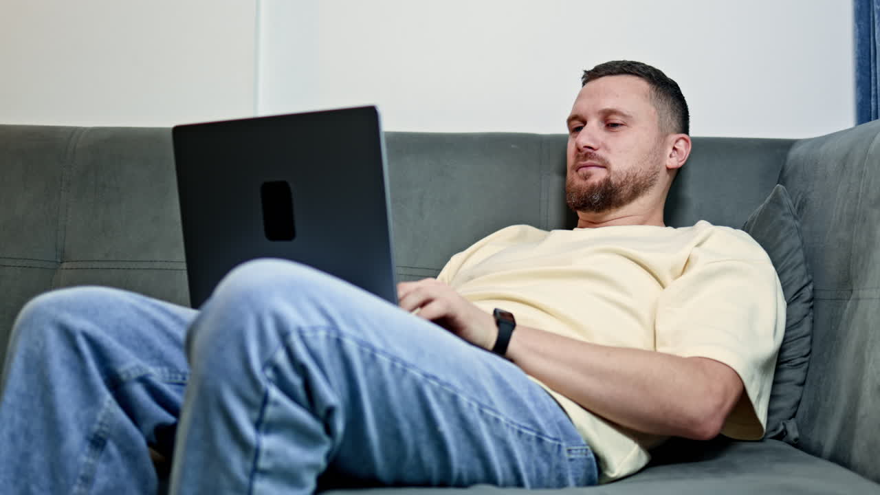 Man works on laptop while sitting on couch in a living room during the day, focusing on tasks and typing in a comfortable position with relaxed posture