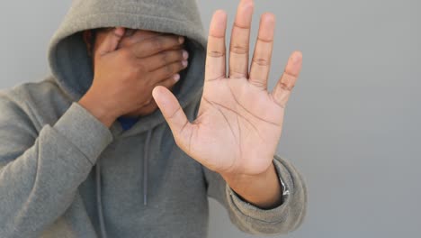 a man wearing a gray hoodie holds up his hand, palm out, in a stop gesture, while covering his face with his other hand.