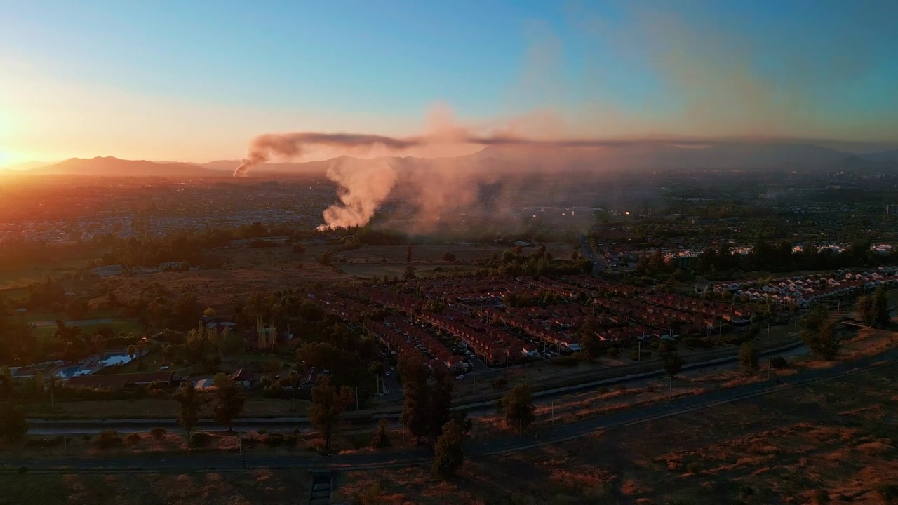 Ampia veduta aerea su un denso sobborgo residenziale al tramonto con drammatici pennacchi di fumo che si innalzano nel cielo arancione e sagome di montagne che brillano all'orizzonte al crepuscolo