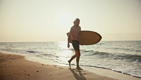 a blond man with a beard with a bare torso and in gray shorts carries a wooden-colored surfboard and walks along the sandy seashore at sunrise in the morning