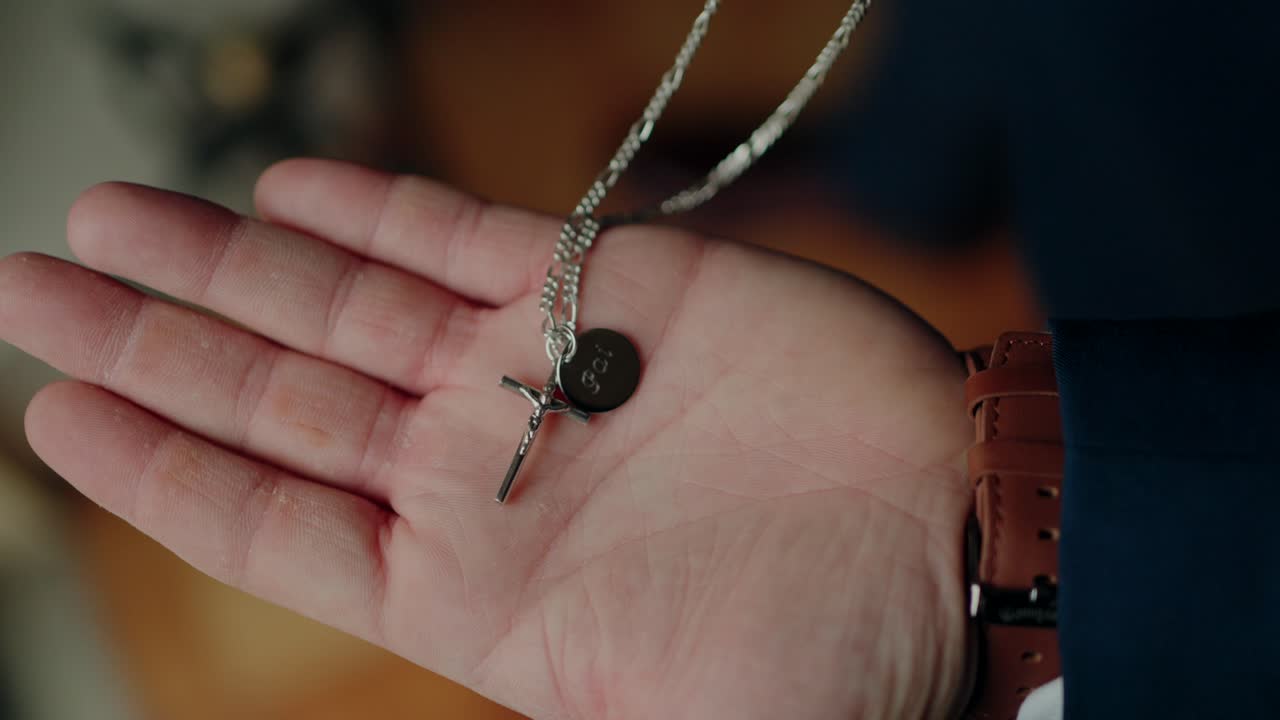 Hand holding a silver cross necklace with a round black charm showing 'G' detail