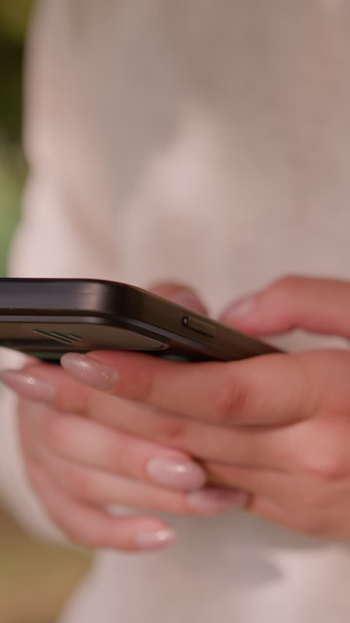 Close-up of individual's hand typing on smartphone with polished nails against softly blurred natural greenery background and unclear figures in the surrounding