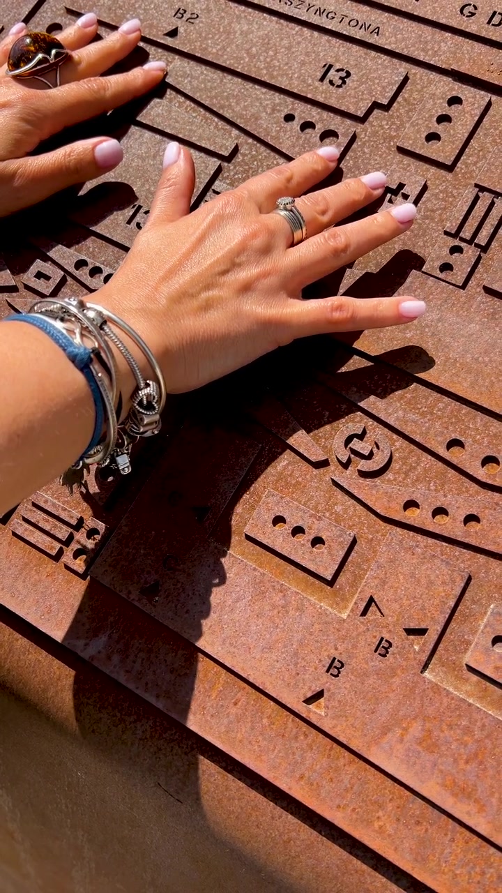 Close-up das mãos de uma mulher explorando um mapa tátil de metal enferrujado com braille e símbolos em relevo. Conceito de design inclusivo e acessibilidade em espaços públicos.