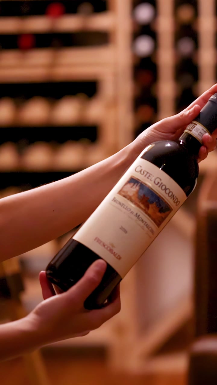 Una persona examina detenidamente una botella de vino tinto en una bodega de vinos con iluminación cálida, girándola para leer la etiqueta. Poca profundidad de campo, cámara estable.