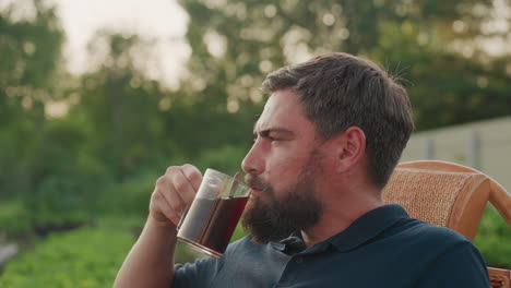 gardener seated in wicker chair at garden edge stirring coffee while sipping hot brew with focused calm expression warm sunset glow backdrop peaceful outdoor break moment