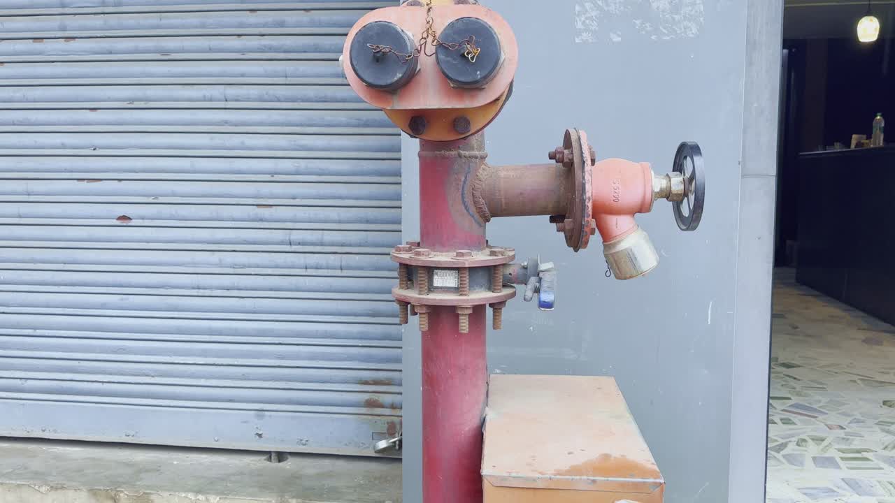 Tilt-up shot of a red industrial fire hydrant pipe with valve and twin outlets, located outside a building near a closed shutter
