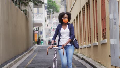 African-american-woman-wearing-face-mask-carrying-bicycle-in-street