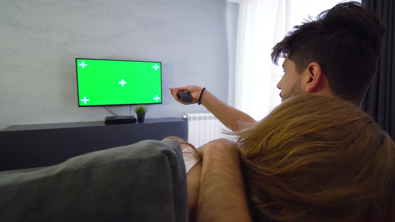 Par ser tv med grøn skærm. Dreng trykker på fjernbetjeningen. Pige krammer dreng