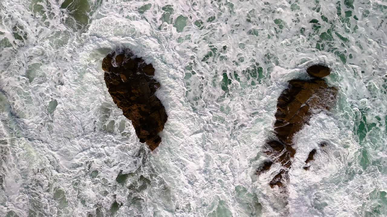 Slow motion, aerial shot of powerful waves with white caps crashing on a set of sea rocks and colourful, turquoise waters