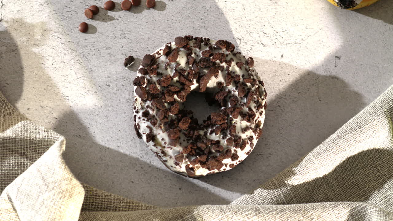 A hand with pink nails grabs a cookie crumble donut topped with chocolate bits from a plate. The scene is warmly lit by sunlight, with a fabric napkin and scattered chocolate drops adding cozy texture