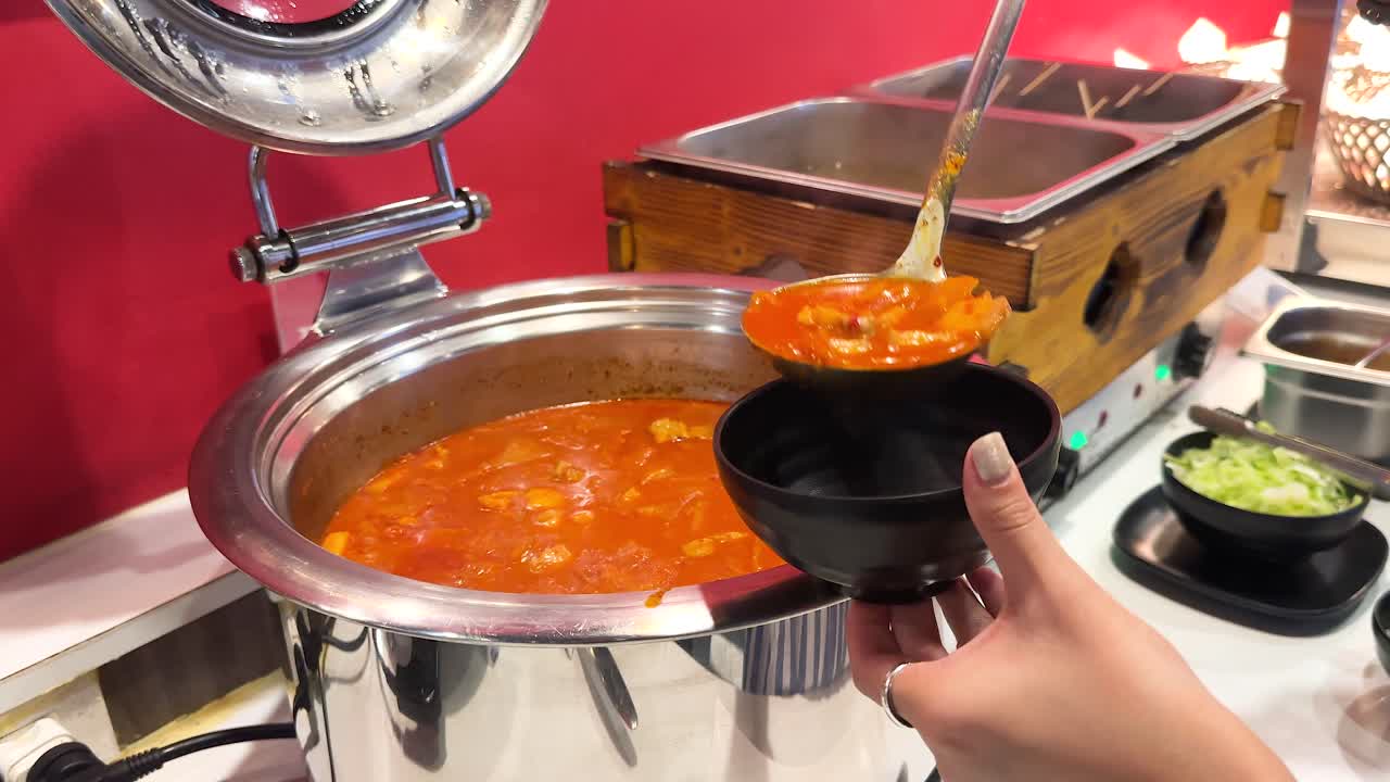 A person ladles spicy kimchi soup into a bowl at a buffet in a vibrant setting