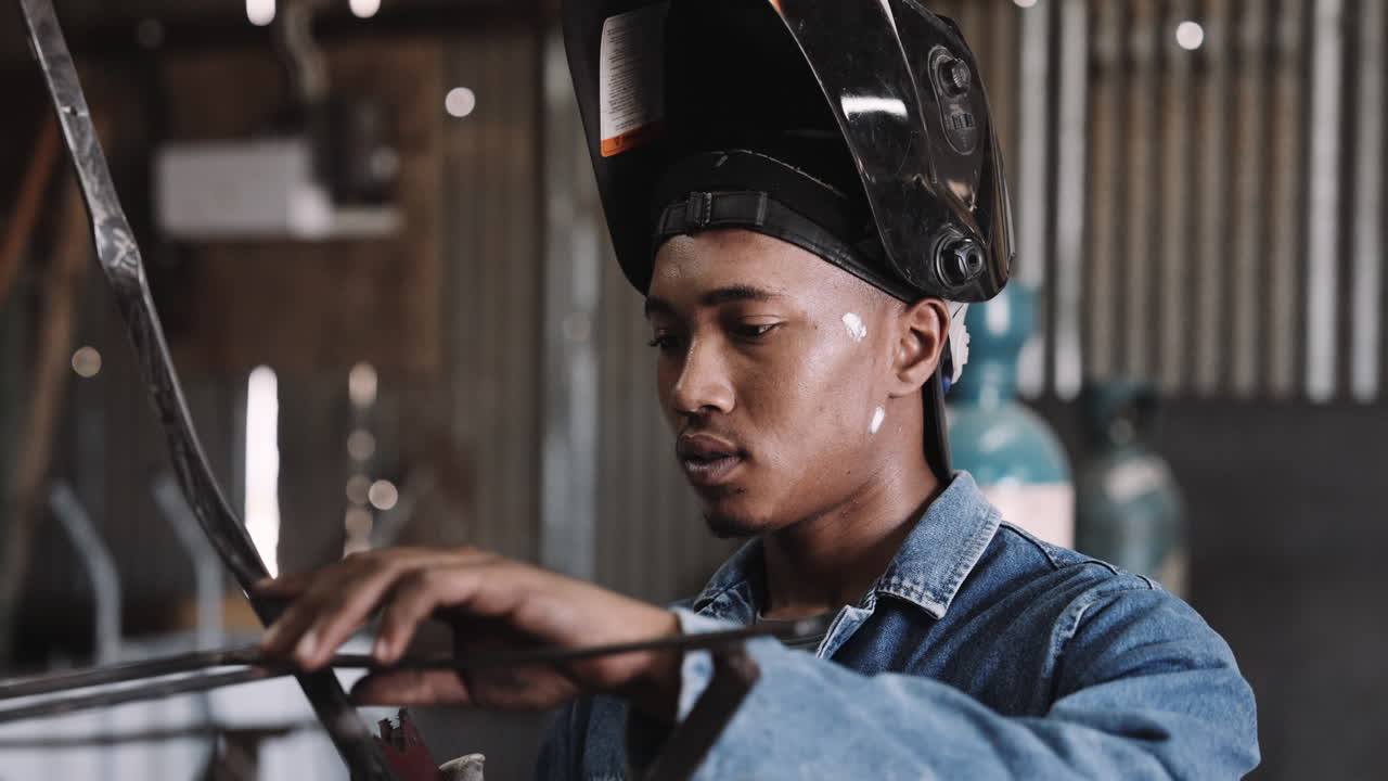 젊은 흑인 남성이 작업실에서 용접 장비와 함께 철조 구조에서 느린 동작을하고 있습니다.