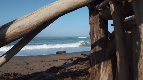 Slow-cinematic-aerial-drone-footage-of-a-fly-through-through-a-wooden-driftwood-tipi-at-a-sandy-beach-at-the-seaside-near-Alberese-in-the-iconic-Maremma-nature-park-in-Tuscany,-Italy,-with-blue-waves