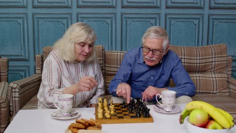 Sonriente-Pareja-De-Ancianos-Abuelo-Abuela-Descansando-En-El-Sofá-Bebiendo-Café,-Jugando-Ajedrez-En-Casa