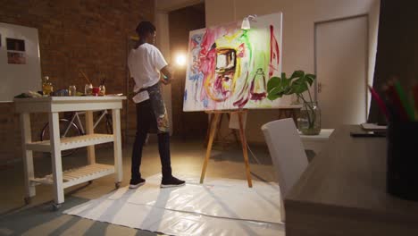 African-american-male-artist-wearing-apron-painting-with-palette-knife-on-canvas-at-art-studio