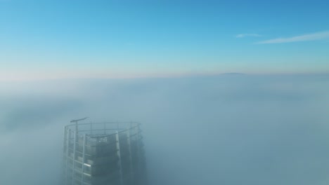 aerial video: drone advances, cruising by a lone skyscraper in thick morning clouds on the left, capturing the cinematic cityscape view