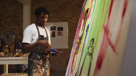 African-american-male-artist-wearing-apron-painting-with-palette-knife-on-canvas-at-art-studio