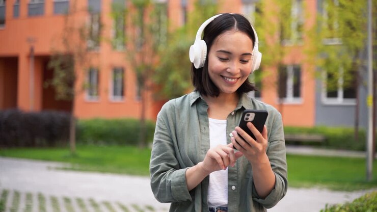 Giovane ragazza giapponese felice che ascolta musica con cuffie wireless mentre cammina all'aperto.