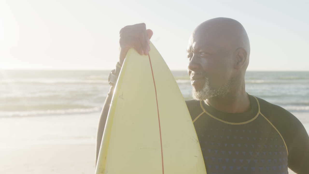 un anciano afroamericano feliz sosteniendo una tabla de surf en la playa, en cámara lenta.