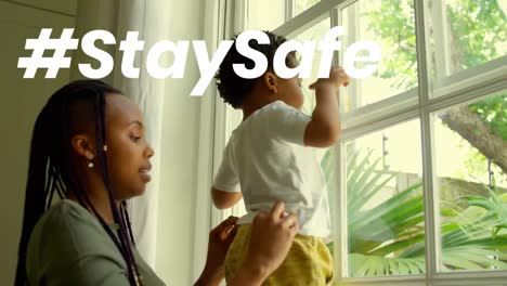 Text-„Bleib-Sicher“-Gegen-Eine-Afroamerikanische-Mutter,-Die-Ihr-Baby-Zu-Hause-Am-Fenster-Hält