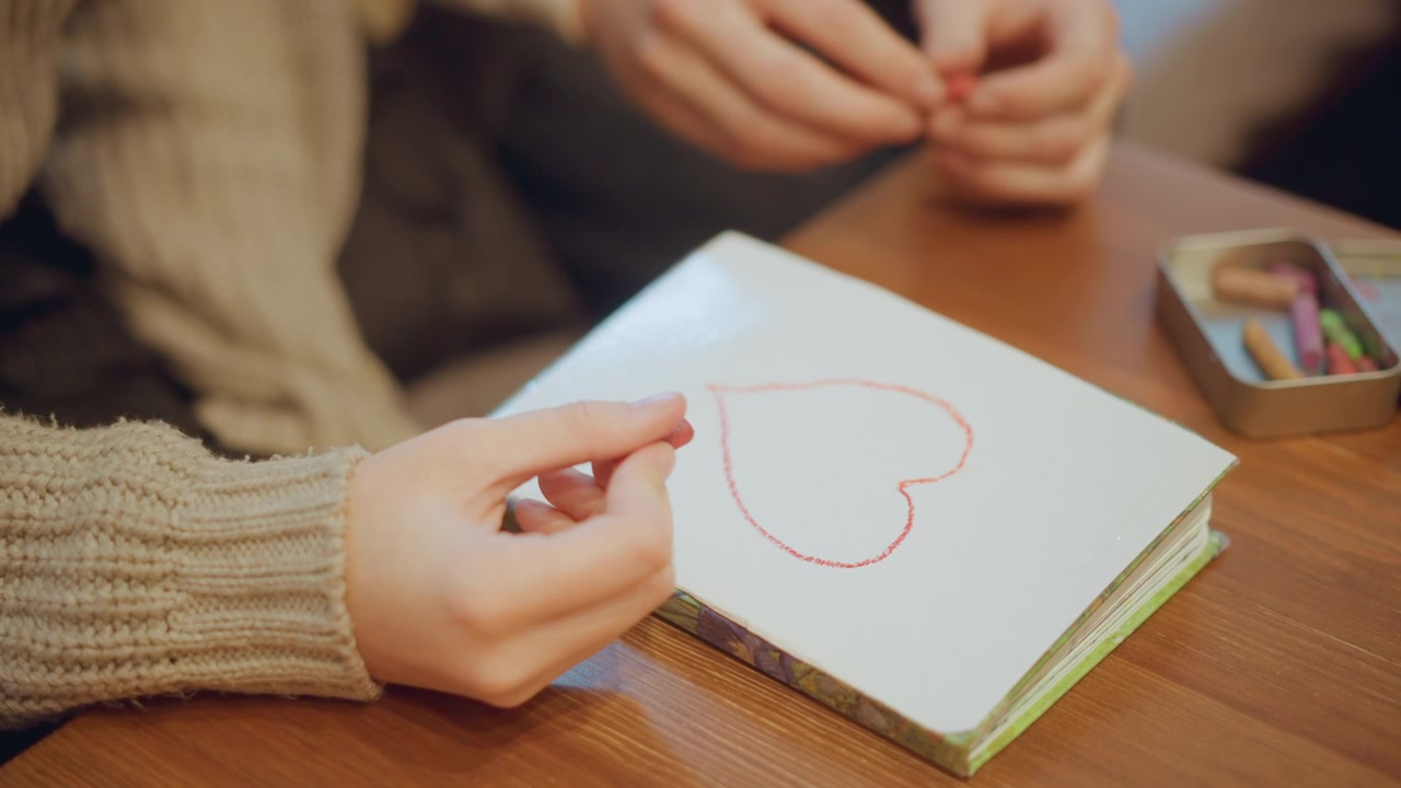 Close up of two people drawing red heart on paper with pastels hands visible. One person holds crayon while another reaches for more colors in soft indoor lighting. small metal tin