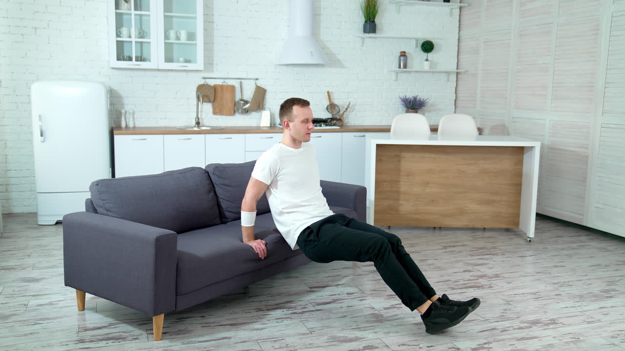 Guy doing workout in the kitchen. Young man doing exercise near the couch at home. Man is training at quarantine.