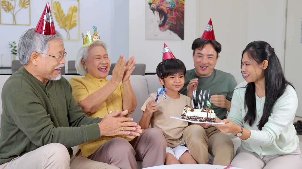 明るいリビングルームで、パーティーハットをかぶった家族が誕生日ケーキを前に、皆で笑顔を見せている。