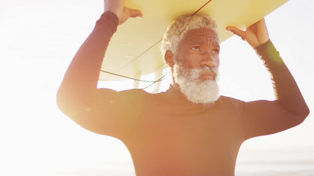 hombre afroamericano mayor caminando con una tabla de surf en una playa soleada