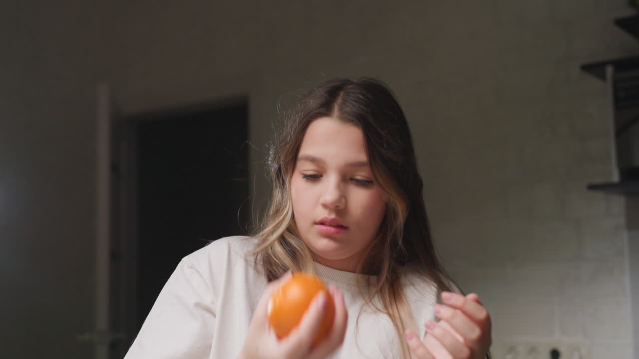 Young girl under ring light in kitchen tossing orange between hands, focused look, soft blur background, calm mood, clean counter, playful motion in arc, skin tones and hair highlights