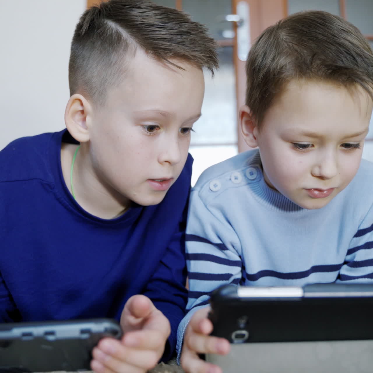 Happy boys playing on wireless gadgets while lying on the carpet at home. Elder boy helps his friend on a digital tablet. Close-up