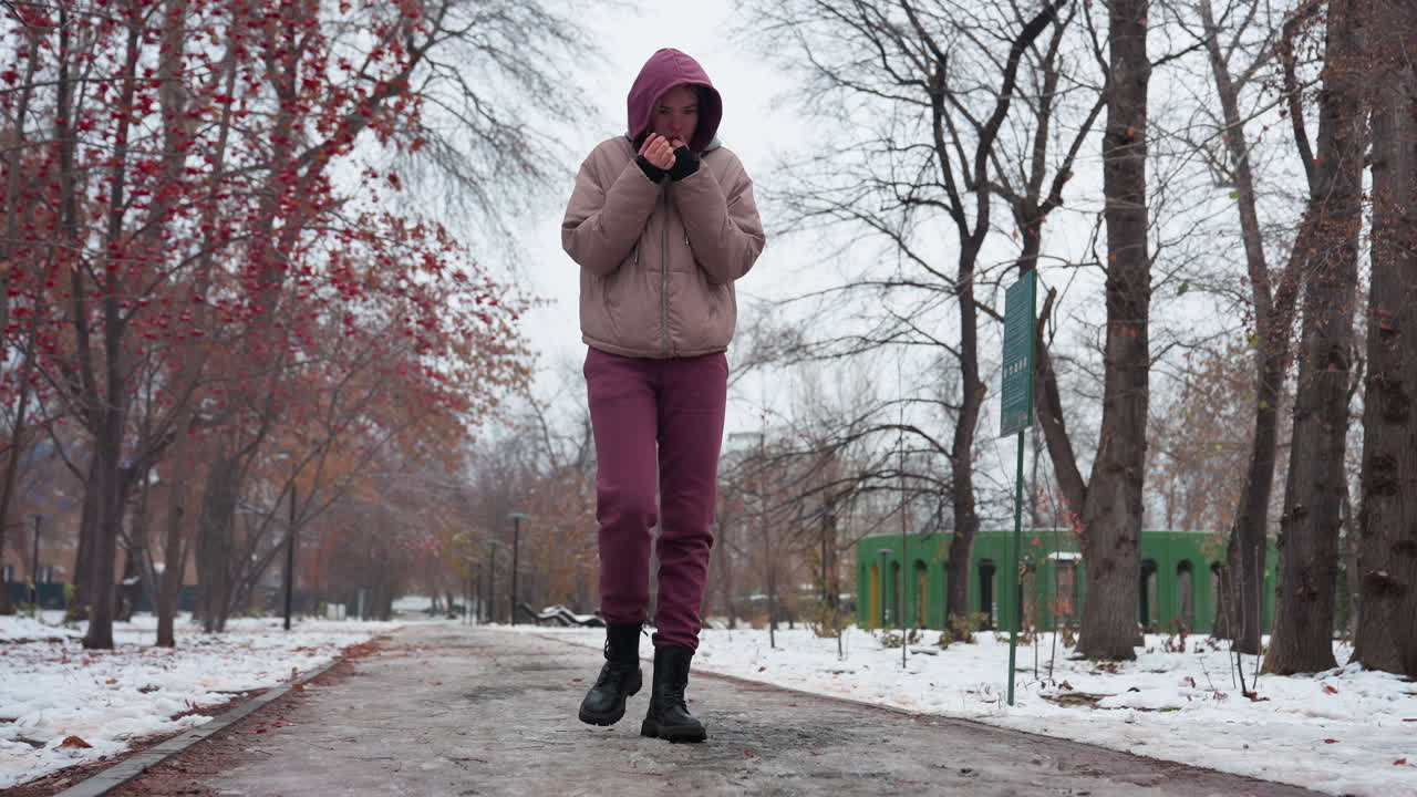 mujer en traje de invierno paseando sola por un parque cubierto de nieve, ajustando la capucha mientras trae la mano cerca de la boca para mantener el calor, árboles desnudos, nieve esparcida y cielo nublado