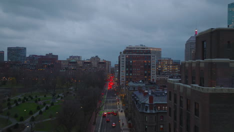 Traffic-on-street-in-urban-borough-at-dusk.-Row-of-brake-lights--on-road-at-traffic-lights.-Forwards-fly-along-public-park.-Boston,-USA