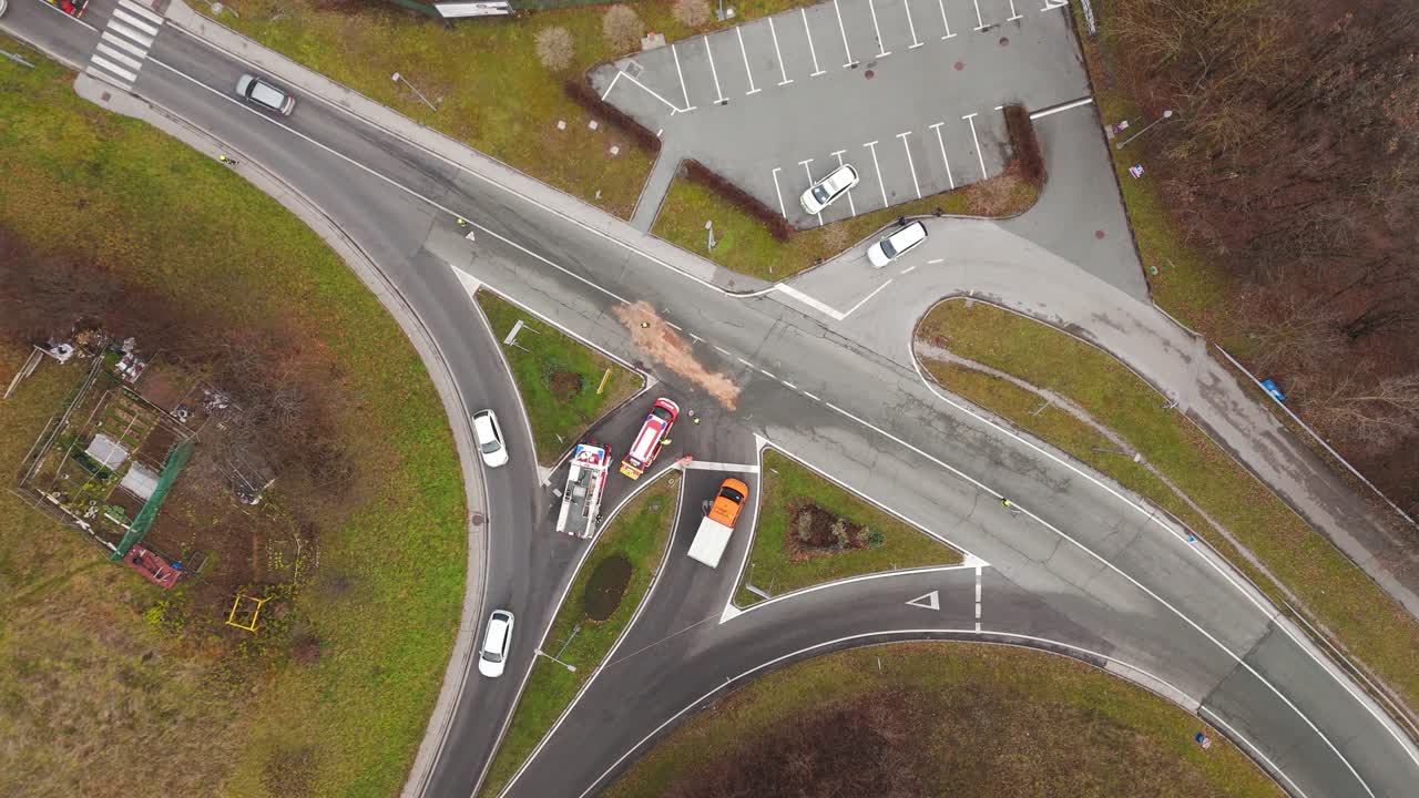 Firefighters cleaning an oil spill after a car crash at a road intersection in Slovenia, with emergency vehicle managing traffic and safety, top-down view