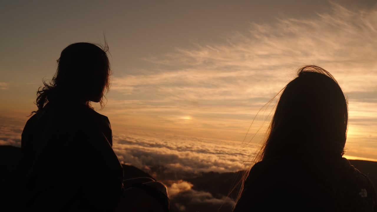 Silhouetted women sit facing a breathtaking sunset above the clouds, immersed in the golden light of the moment.