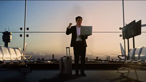 full body of asian businessman with rolling suitcase waits for flight in boarding lounge, looking at a laptop and celebrating, airport terminal with airplane takes off outside the window