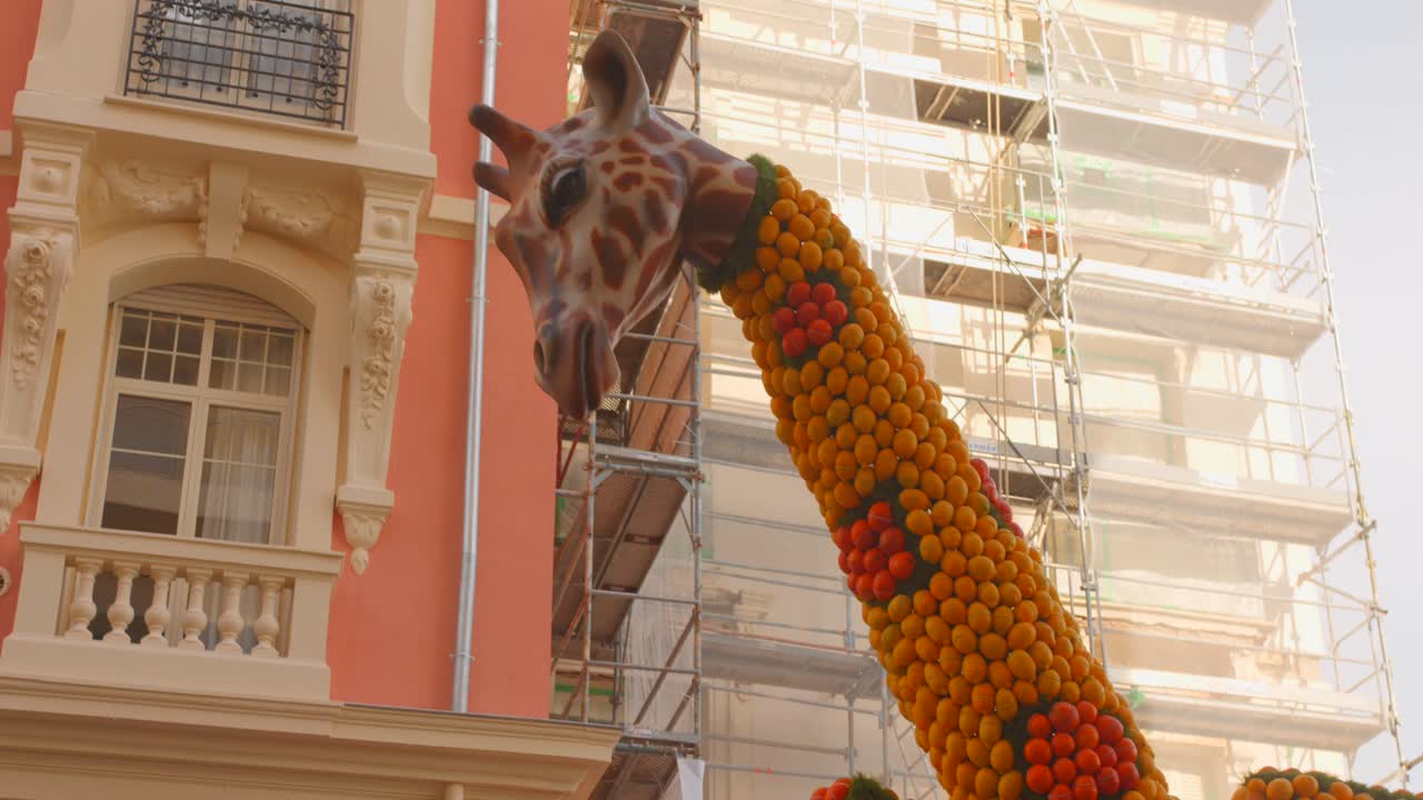 Gran escultura de jirafa hecha de fruta con un edificio de fondo.