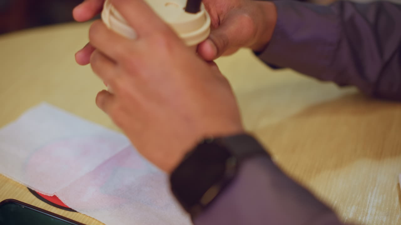 Close-up of hand holding cup beside smartphone on wooden table, with napkin partially covering red circular design. Person wears wristwatch and purple shirt, capturing casual moment in indoor setting