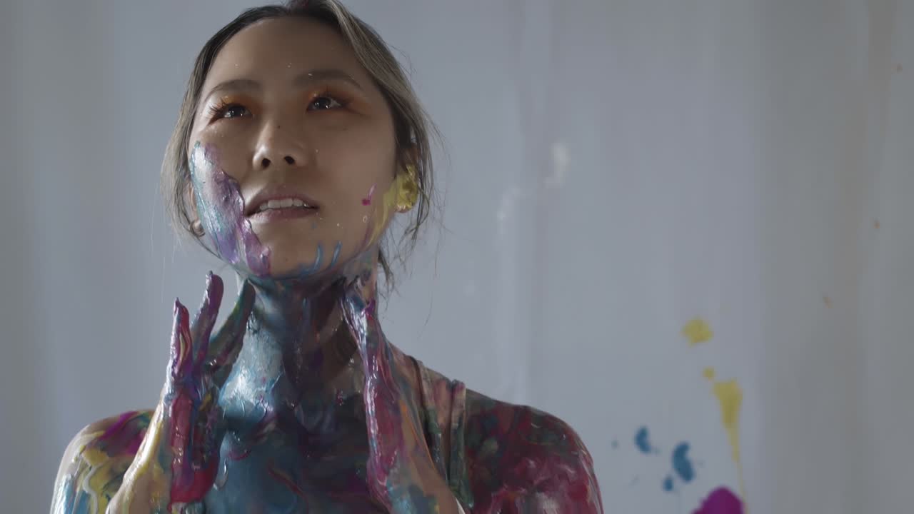Asian girl covered in colorful paint poses in studio, handheld closeup