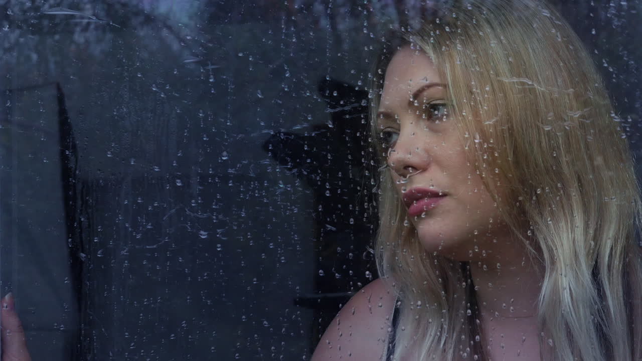 Triste e sola giovane donna bionda che guarda fuori da una finestra sotto la pioggia.
