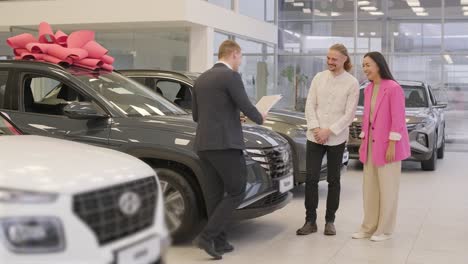 young car salesman showing to young couple new automobile at dealership salon.