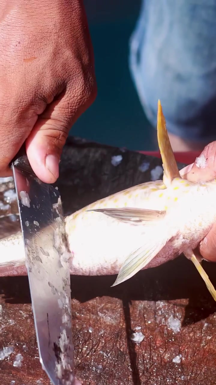 Una persona usa un grande coltello per desquamare un pesce maculato su un tagliere di legno rustico sotto la luce solare naturale intensa, con acqua visibile sullo sfondo