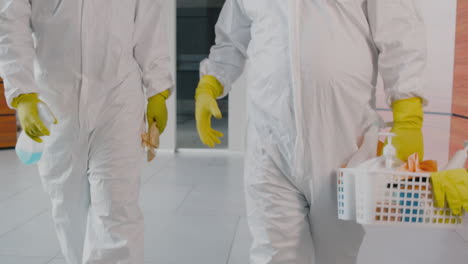 two unrecognizable cleaning men wearing personal protective equipment while walking inside an office building holding cleaning products and rags