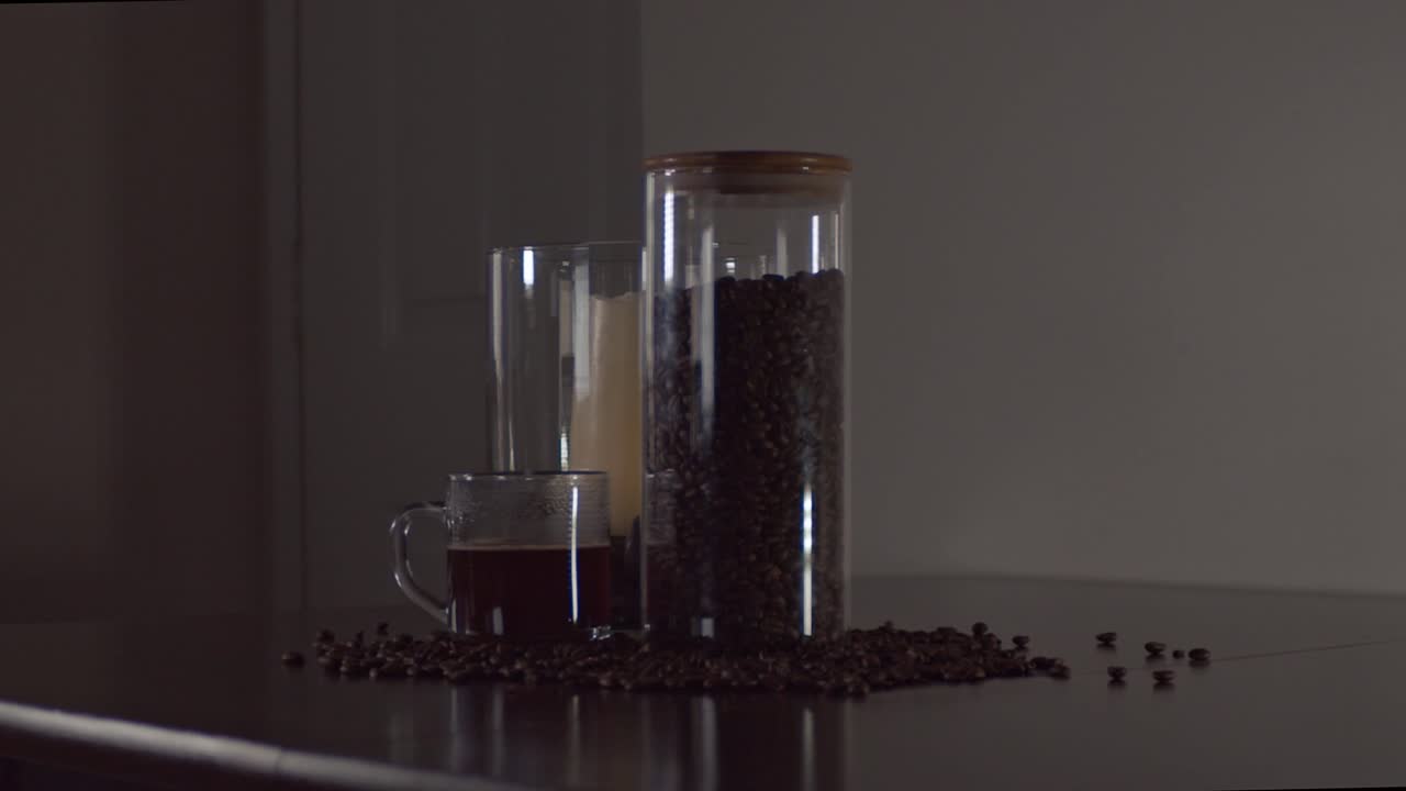 Coffee beans, a glass container, candle centerpiece, and a steaming glass mug of coffee  sit on a table as the camera slides with a wide shot.