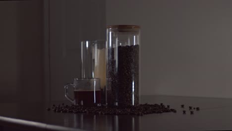 coffee beans, a glass container, candle centerpiece, and a steaming glass mug of coffee  sit on a table as the camera slides with a wide shot.