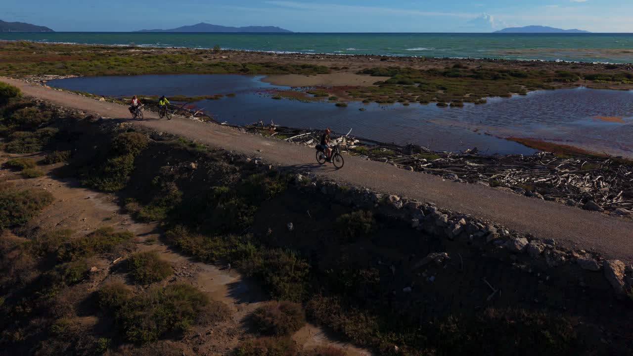 E-bike riders on a coastal trail with sea, forest and islands in Parco Maremma. Group of three aerial