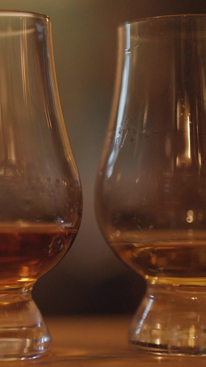 Due bicchieri da whisky con liquido ambrato poggiano su una superficie di legno, illuminati da una luce soffusa e calda. La telecamera esegue una lenta panoramica, creando un'atmosfera rilassata e intima.
