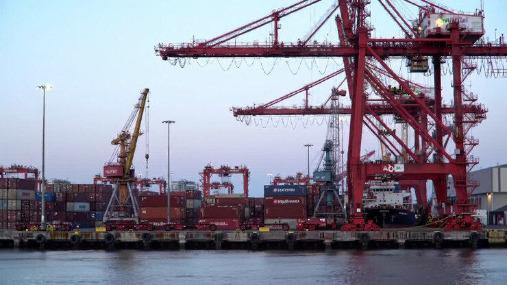 Container cranes lift cargo from vessels as dusk sets in at a busy shipping terminal. Bright lights illuminate the scene, emphasizing the ongoing maritime activities in the port.
