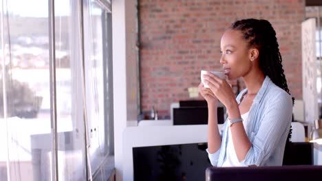 African-american-casual-businesswoman-drinking-coffee-by-window-in-office,-slow-motion,-copy-space